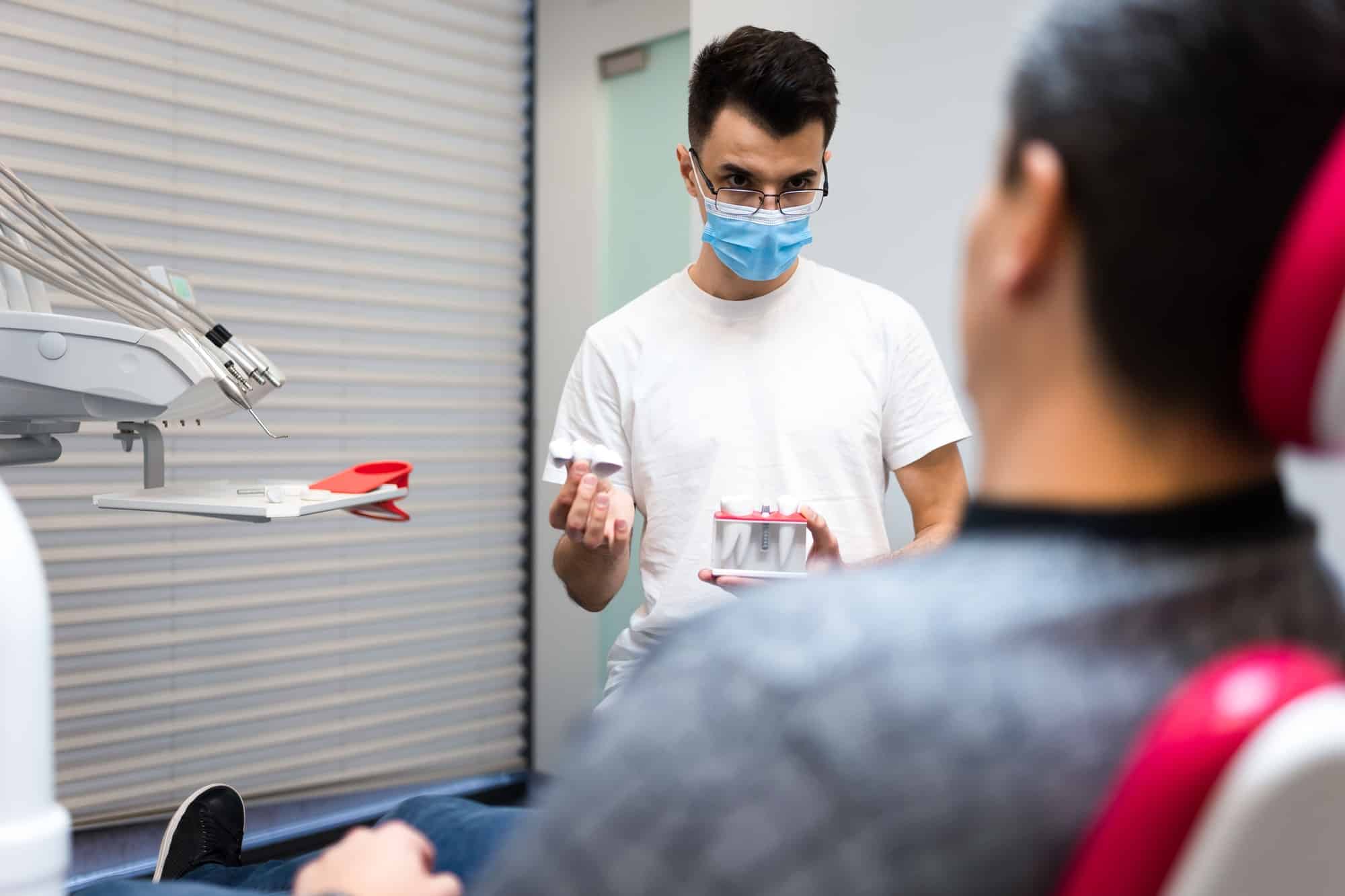 Dental surgeon with dental crown in hands model talks to patient about prosthetics.